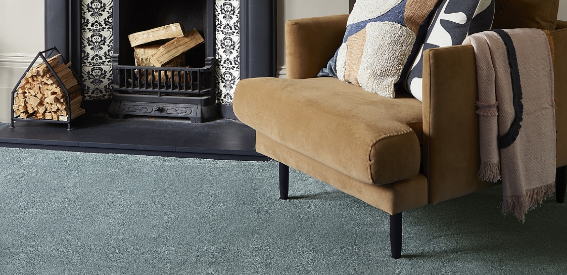 A blue shade of carpet laid in a country style living area with mustard armchair and fireplace.